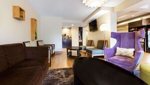Una sala de estar moderna con un sofá marrón, un reposapiés negro, un sillón púrpura y una mesa de centro redonda. Hay un televisor montado en la pared y un estante de madera con un objeto decorativo.