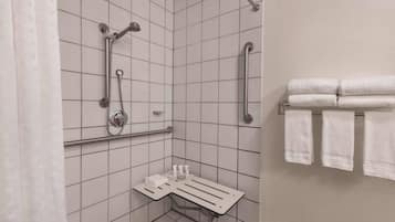 A bathroom with white tiled walls, a shower with grab bars, a towel rack with folded towels, and a shelf with toiletries.