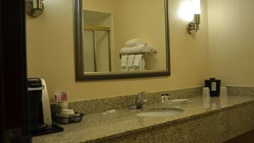 A bathroom with a granite countertop, a sink, a mirror, a towel rack with folded towels, a coffee maker, and toiletries.
