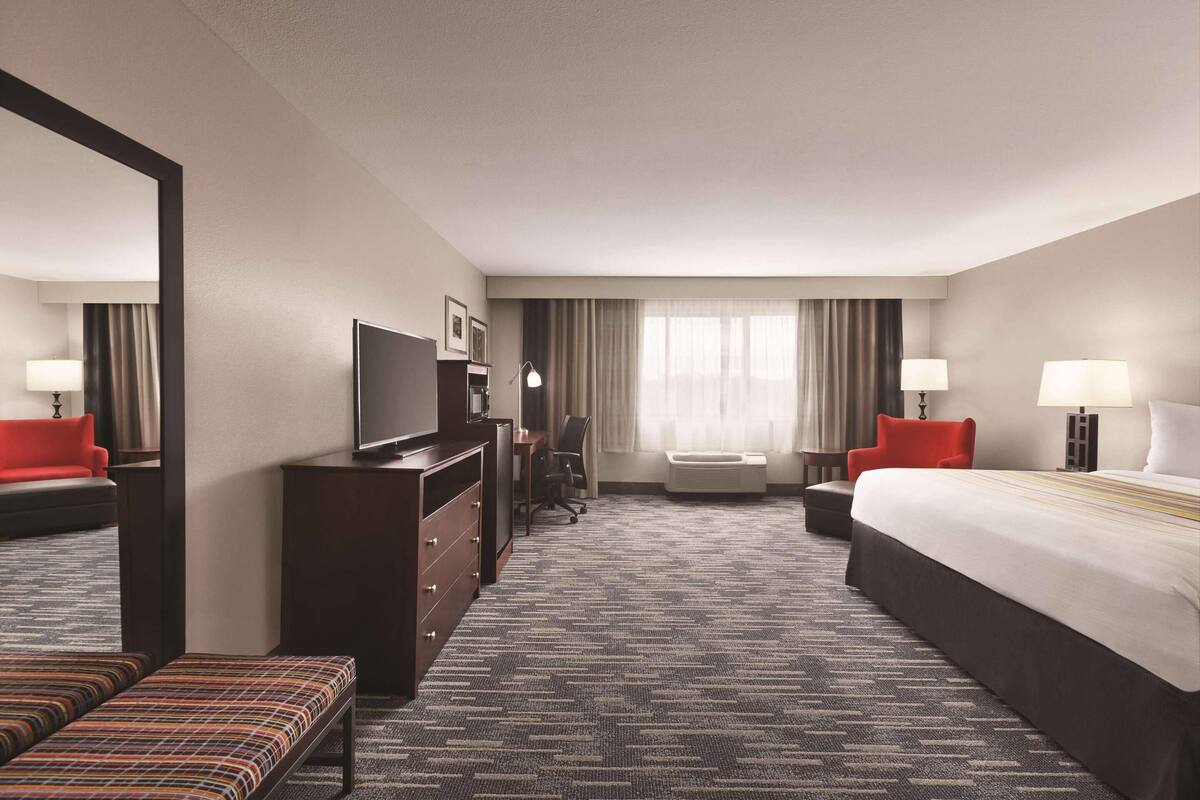 A hotel room with a large bed, a desk with a chair, a television, a dresser, and a bench.