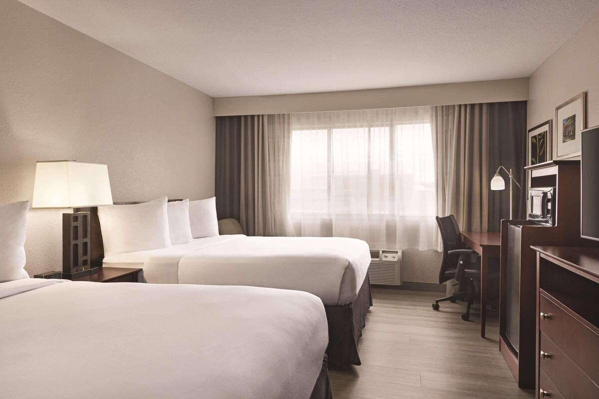 A hotel room with two beds, a desk, a chair, a TV, and a lamp.