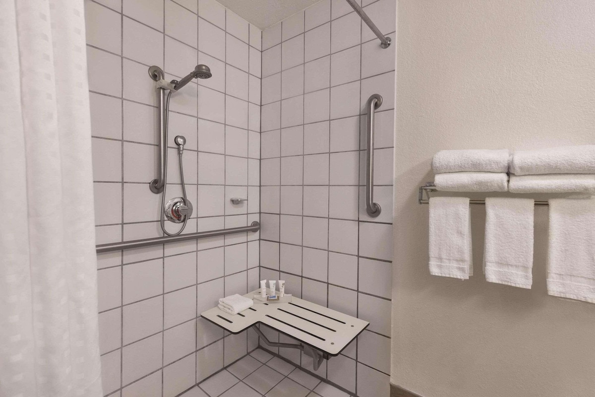 A small, tiled bathroom with a shower, grab bars, a towel rack, and a shelf with toiletries.