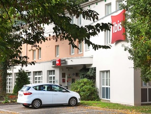 A white car parked in front of a building with a red 'ibis' sign, indicating a hotel or similar establishment.