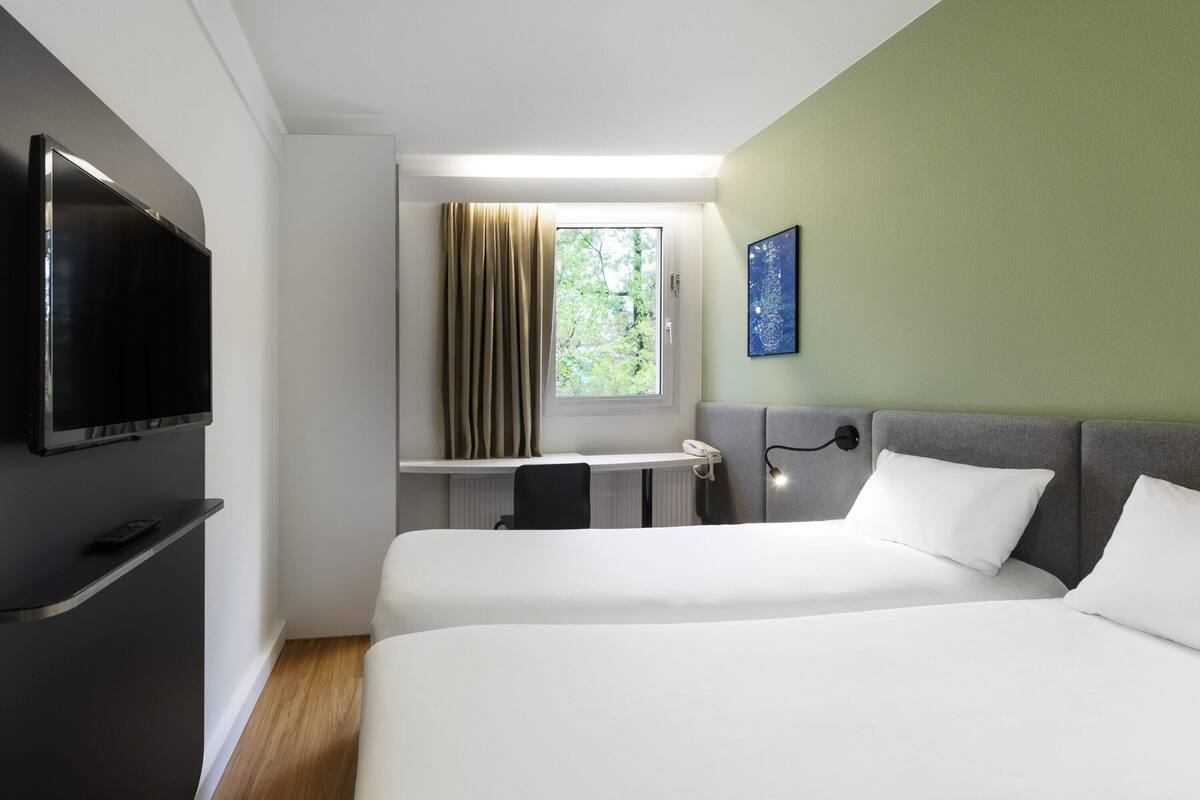 A hotel room with two beds, a TV, a desk with a chair, a window with curtains, and a painting on the wall.