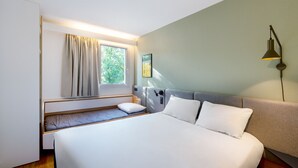 A bedroom with a bed, bedside table, lamp, window with curtains, and a view of greenery outside. - ibis Nancy Brabois (Vandœuvre-lès-Nancy)