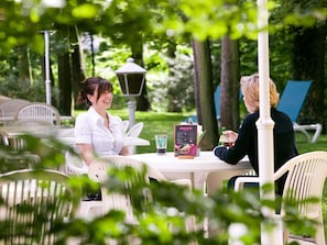 Deux personnes sont assises Ă une table Ă lâextĂ©rieur, devant un menu et des boissons, entourĂ©es de verdure et de chaises blanches.
