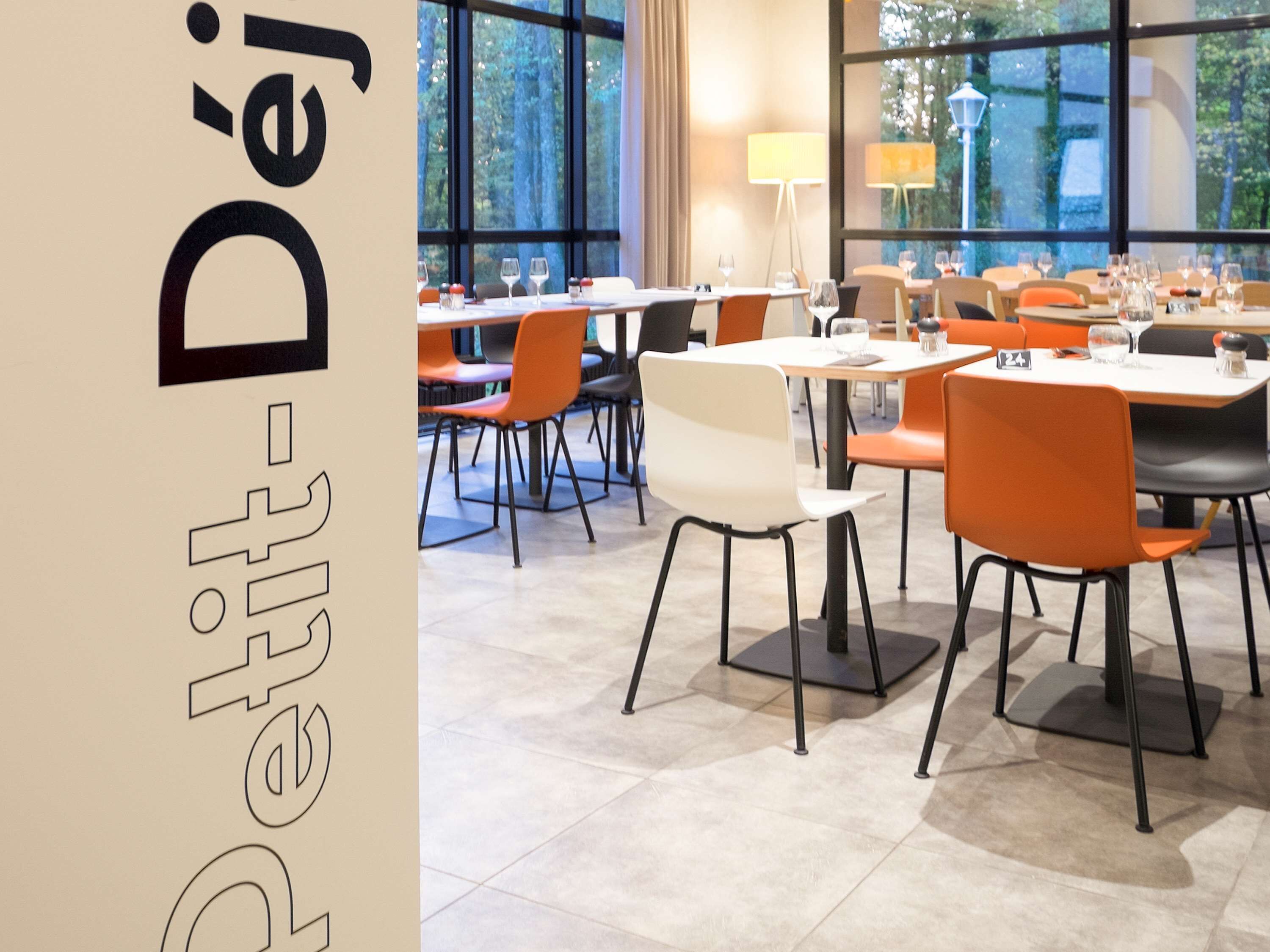 A modern dining area with tables and chairs, large windows with a view of trees, and a lamp on the wall.