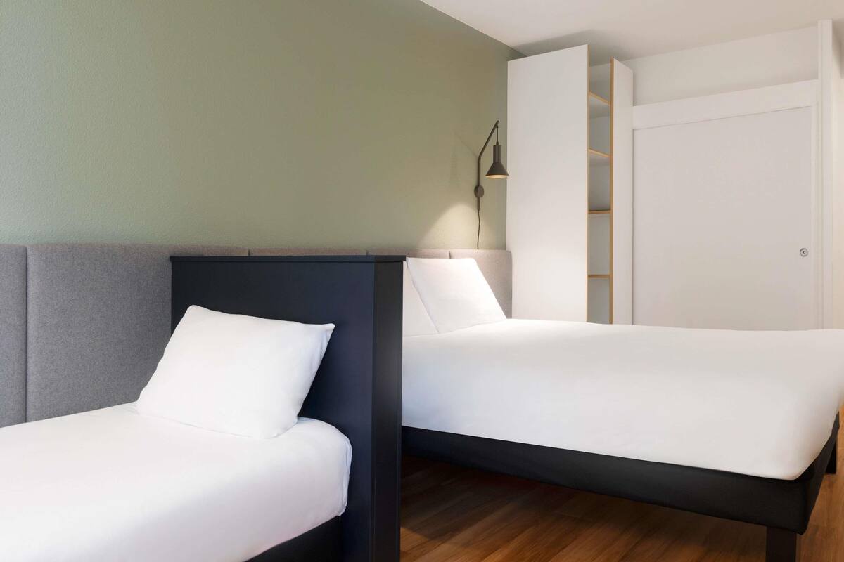 A modern hotel room with two single beds, a wardrobe, and a bedside lamp.