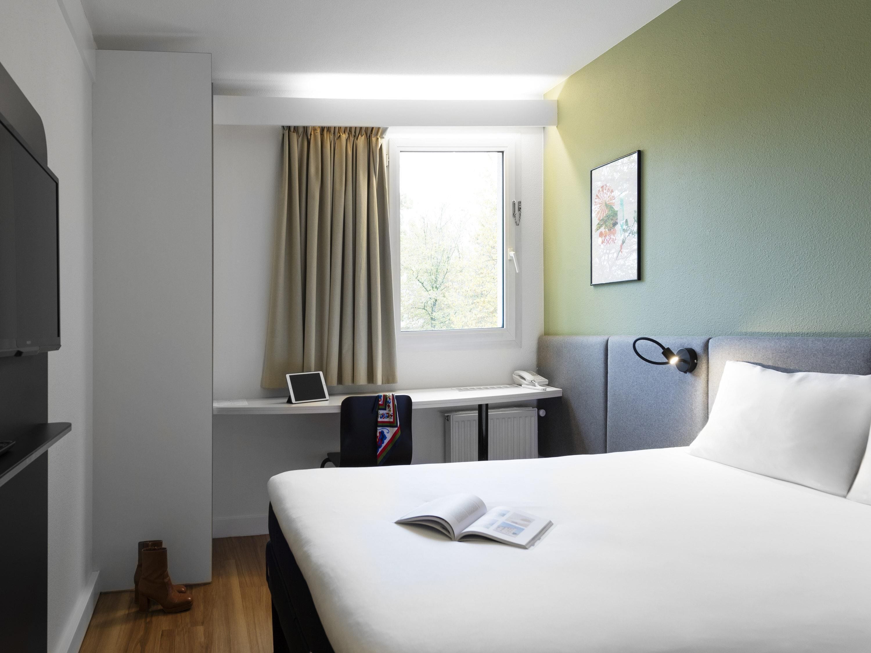 Una habitación de hotel con cama, escritorio, computadora portátil, teléfono y ventana con cortinas.