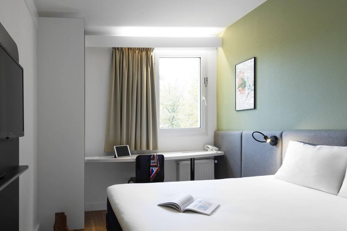 A hotel room with a bed, a desk with a laptop, a telephone, and a window with curtains.