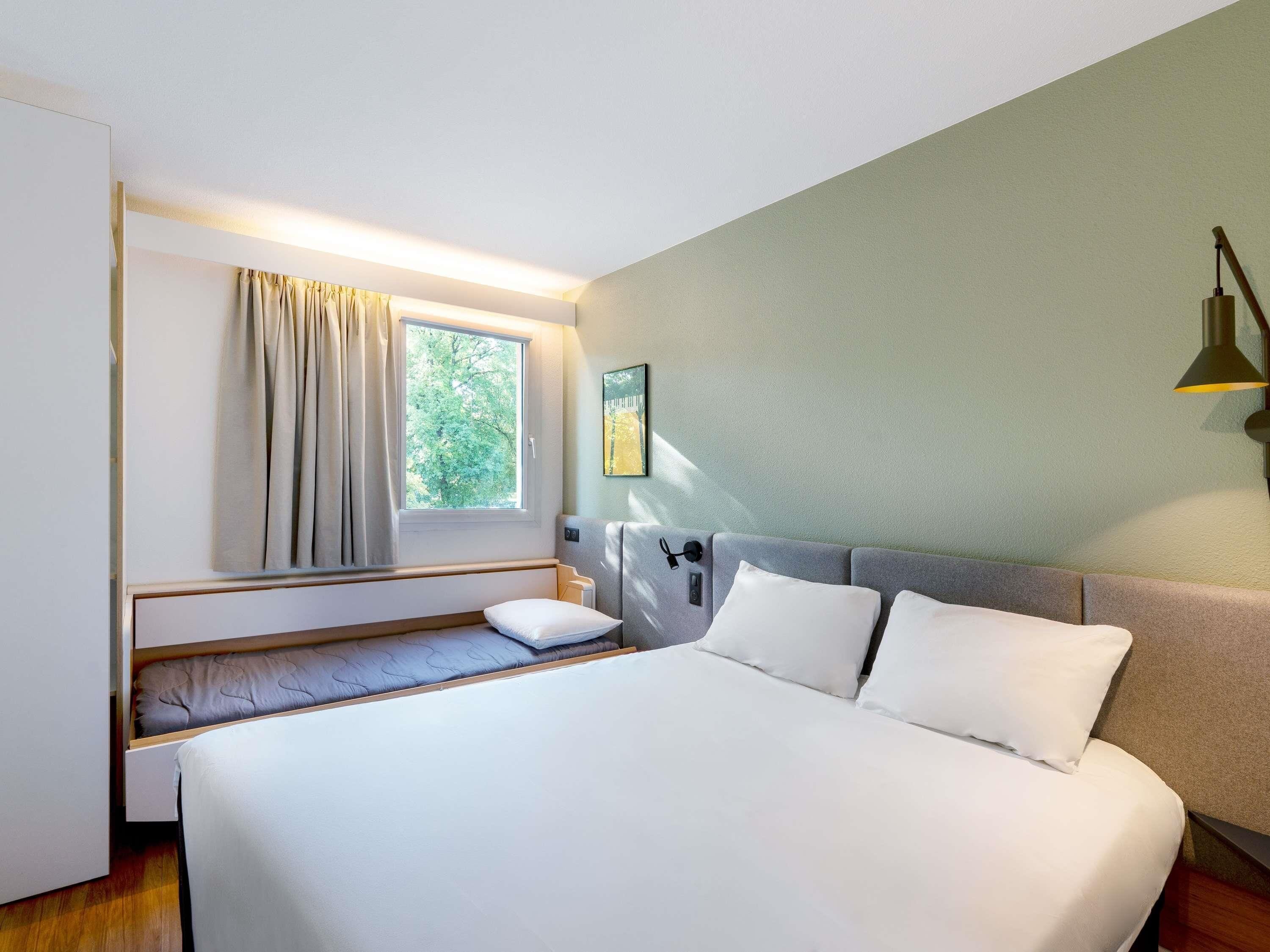 Habitación de hotel pequeña con cama, ventana con cortinas, mesita de noche y lámpara de pared.