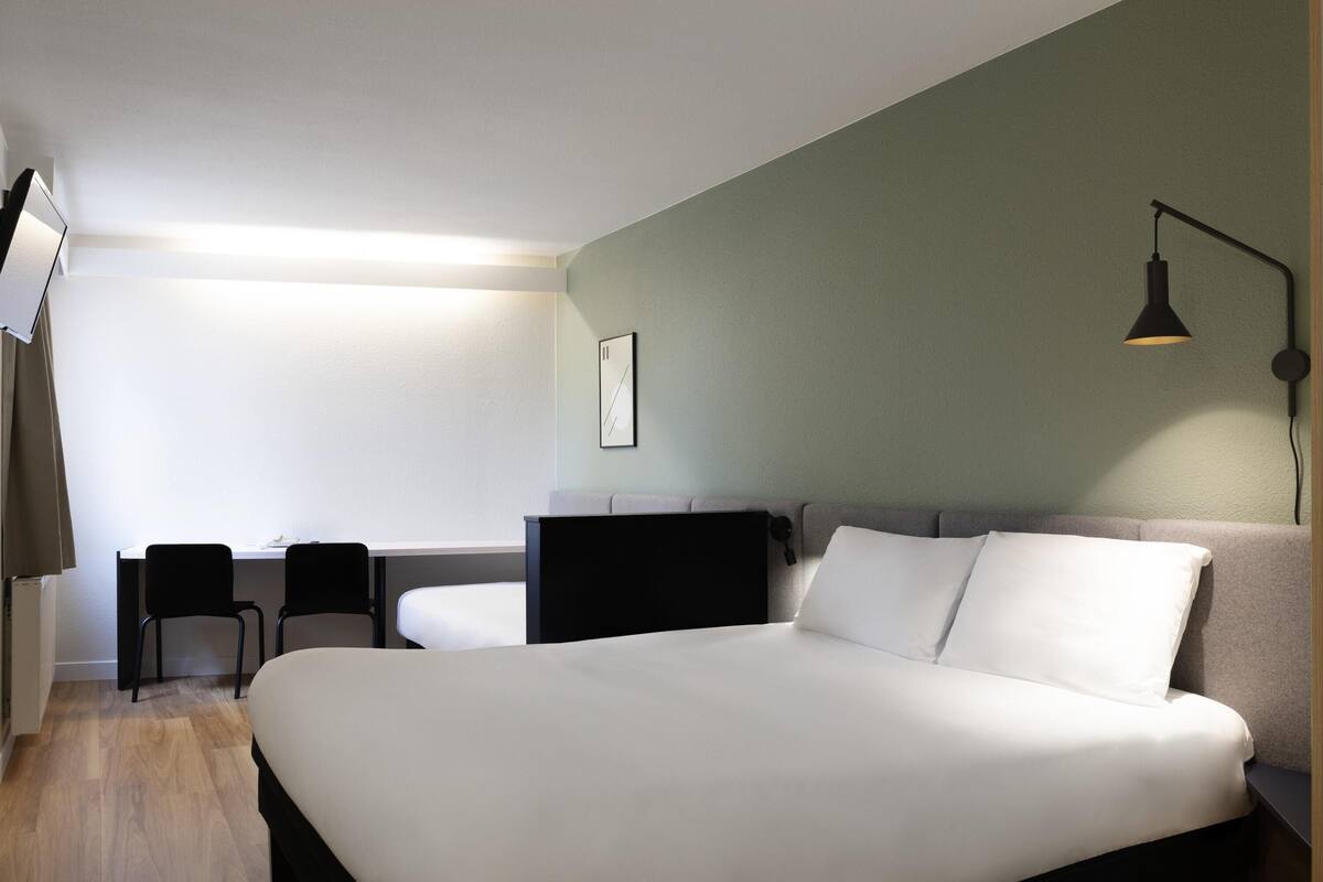 A bedroom with a large bed, a desk, two chairs, a wall-mounted lamp, and a framed picture.