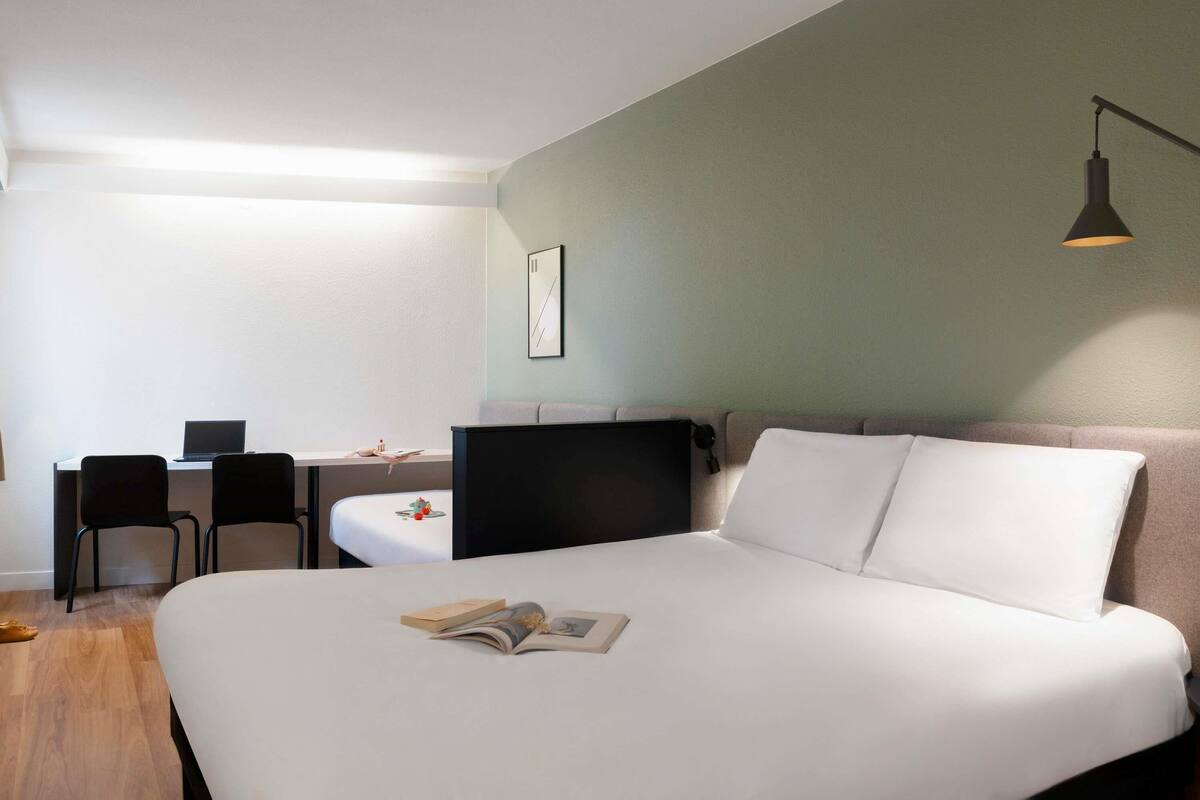 A modern hotel room with a large bed, a desk with a laptop, and a small table with a lamp.