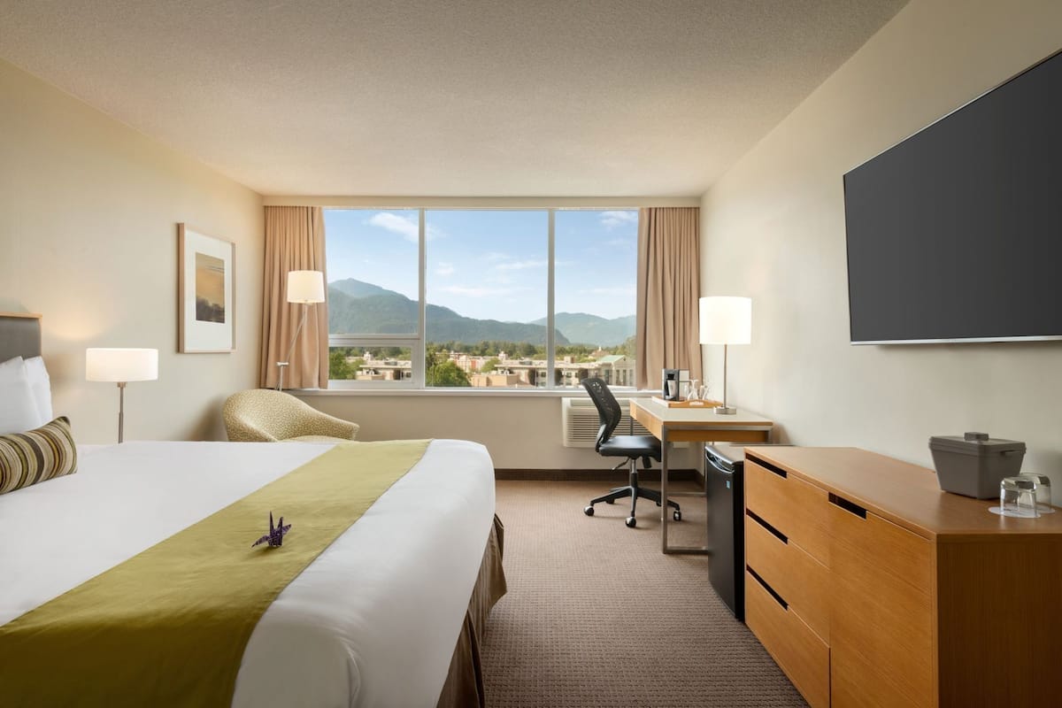 A hotel room with a bed, desk, chair, and a large window with a mountain view.