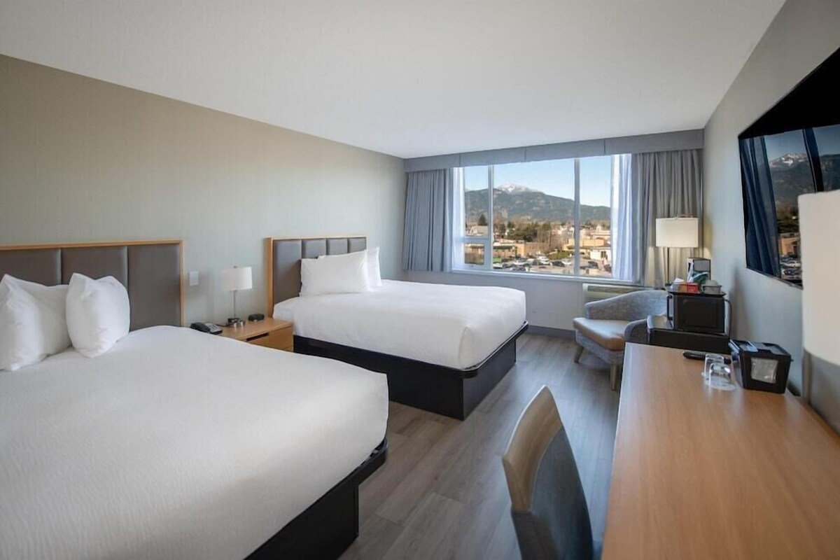 Habitación de hotel con dos camas, un escritorio, una silla, un televisor y una ventana con vistas a la ciudad.