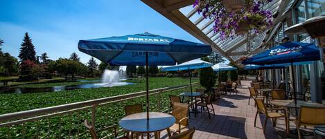 An outdoor patio area with tables and chairs, umbrellas, a fountain, and a view of a pond with water lilies.