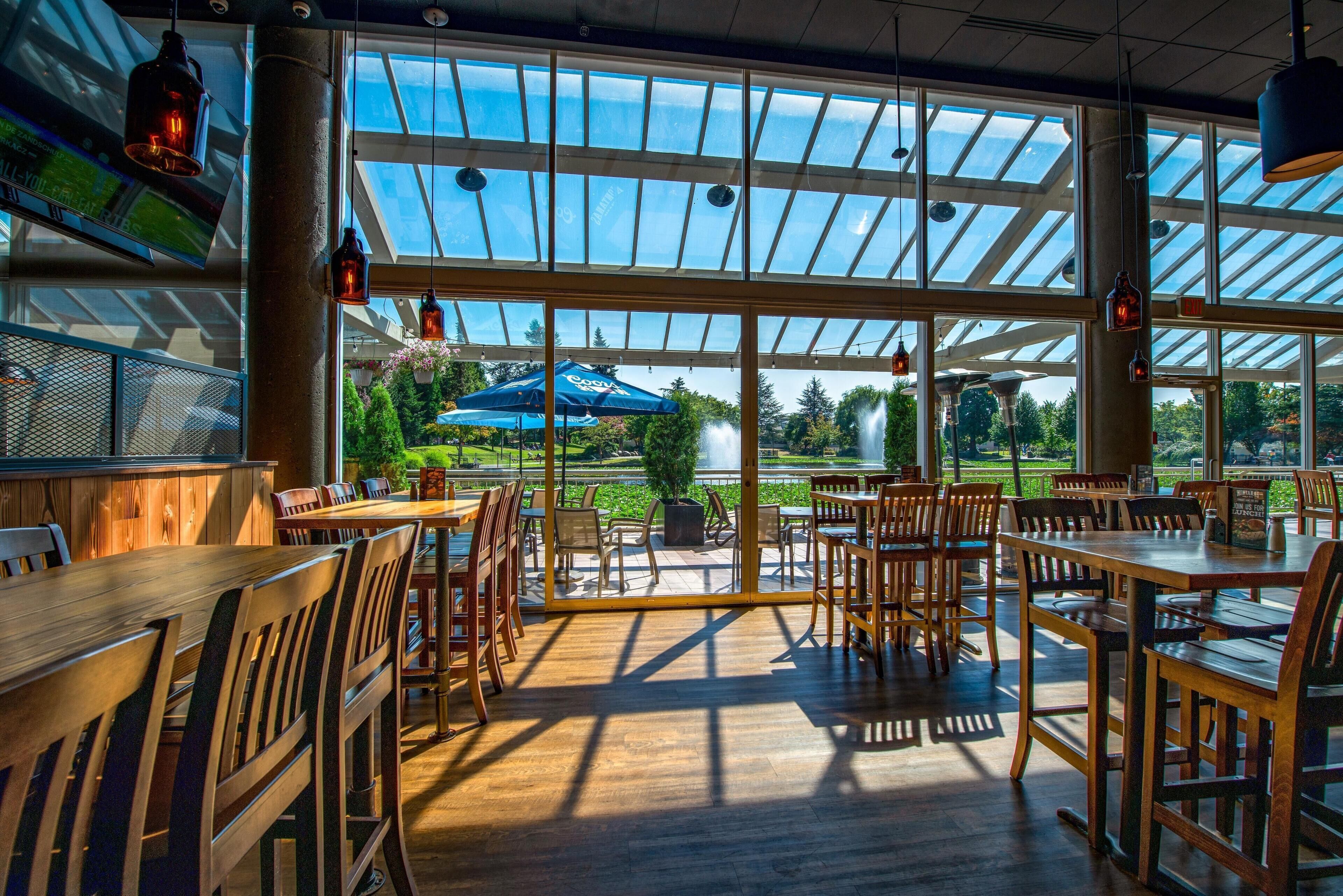 Un restaurant avec des tables et des chaises en bois, de grandes fenêtres donnant sur un paysage verdoyant et des lampes suspendues.