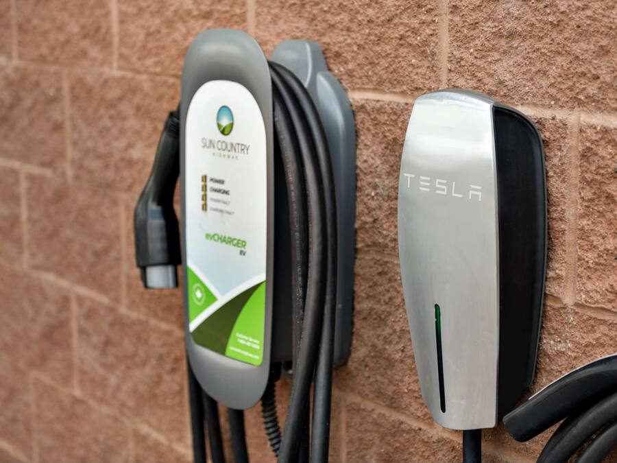 Dos estaciones de carga para vehículos eléctricos, una con la marca 'Sun Country' y la otra 'Tesla', fijadas a una pared de ladrillo.