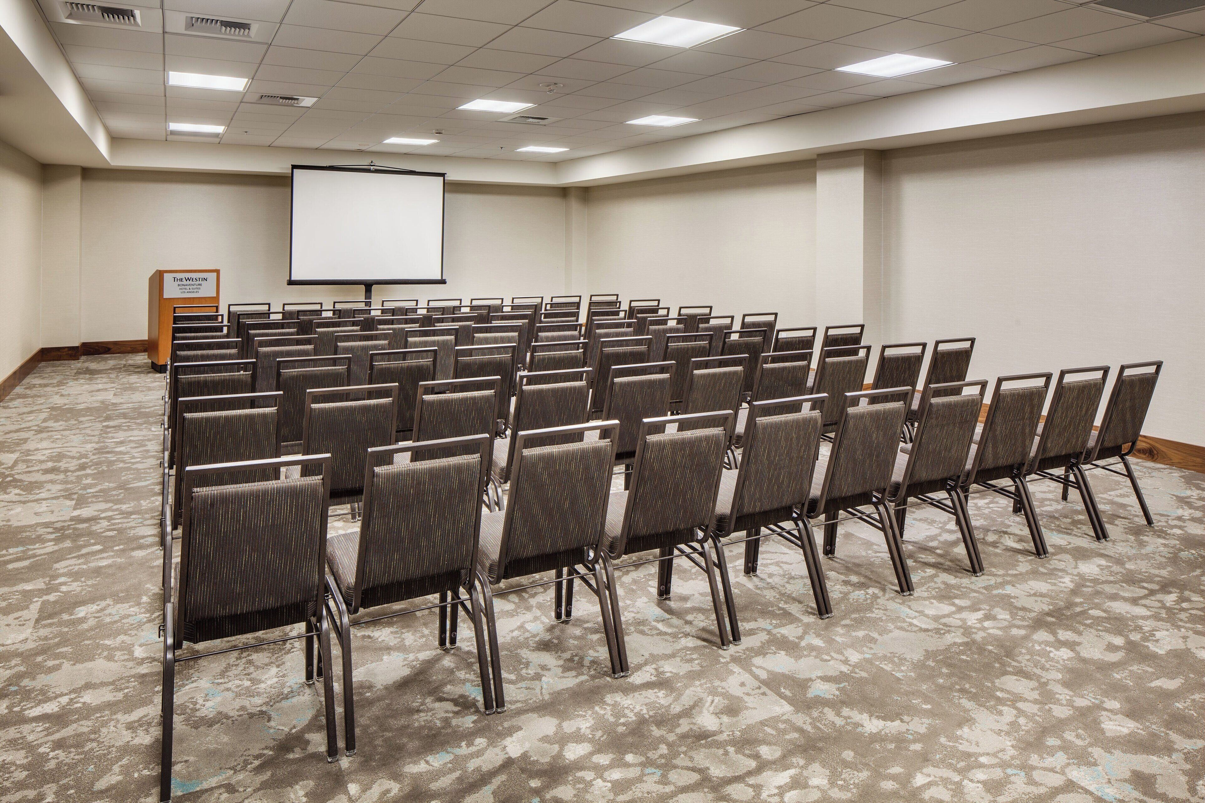 Un salón de conferencias con filas de sillas, una pantalla para proyector y un atril.