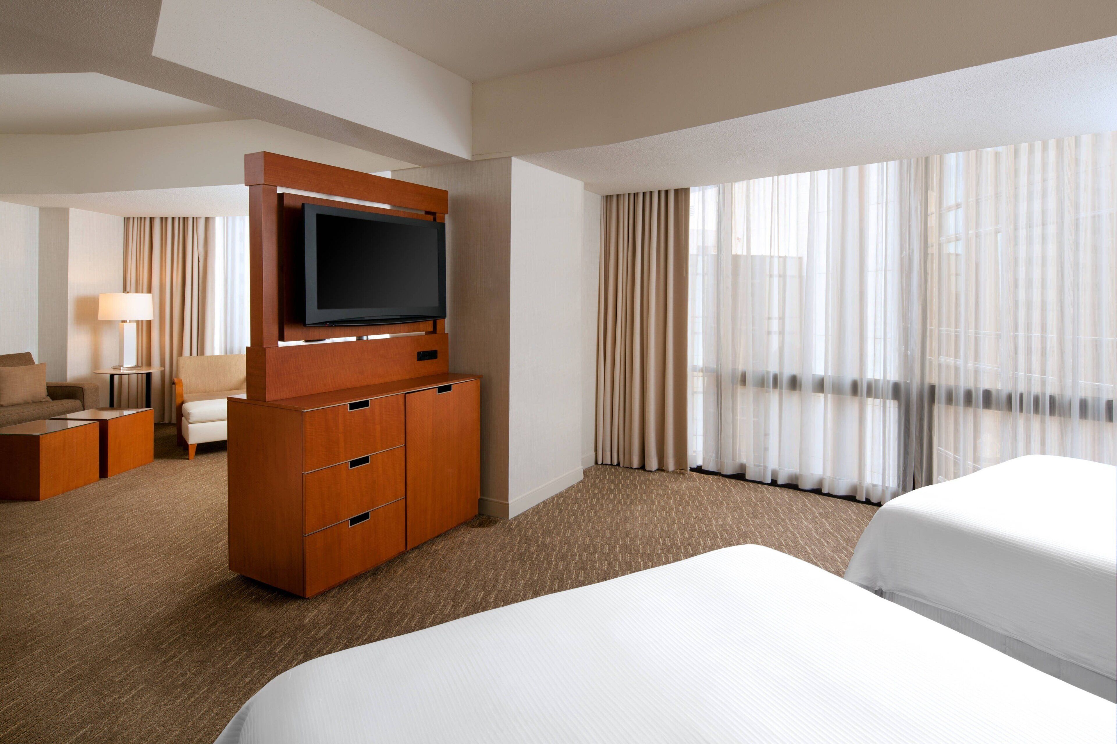 A hotel room with a large bed, a TV mounted on a wooden cabinet, a desk with a lamp, and a view of the city through sheer curtains.