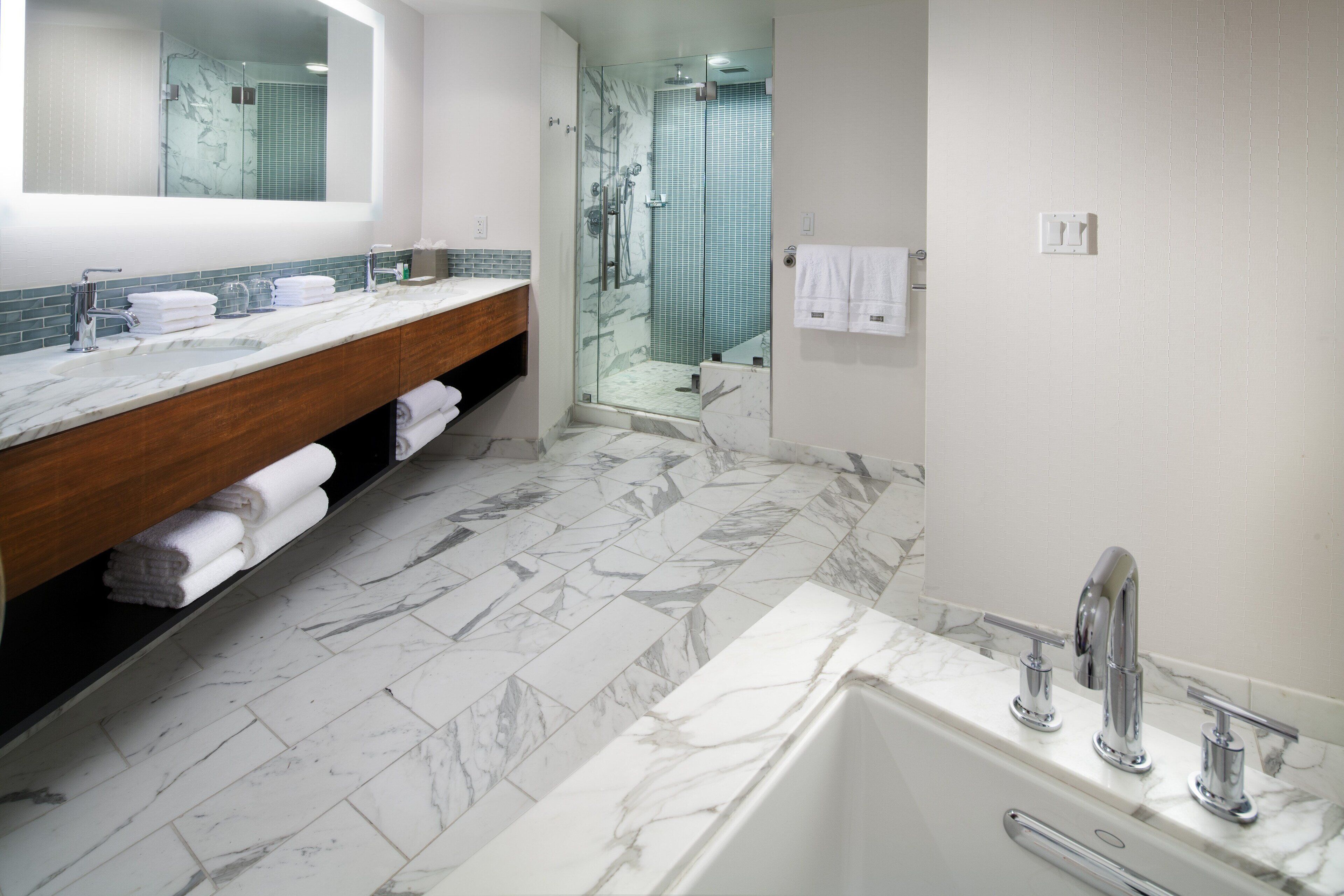 Un baño moderno con piso de mármol, una bañera grande, un tocador de madera con espejo y una ducha con cerramiento de vidrio.