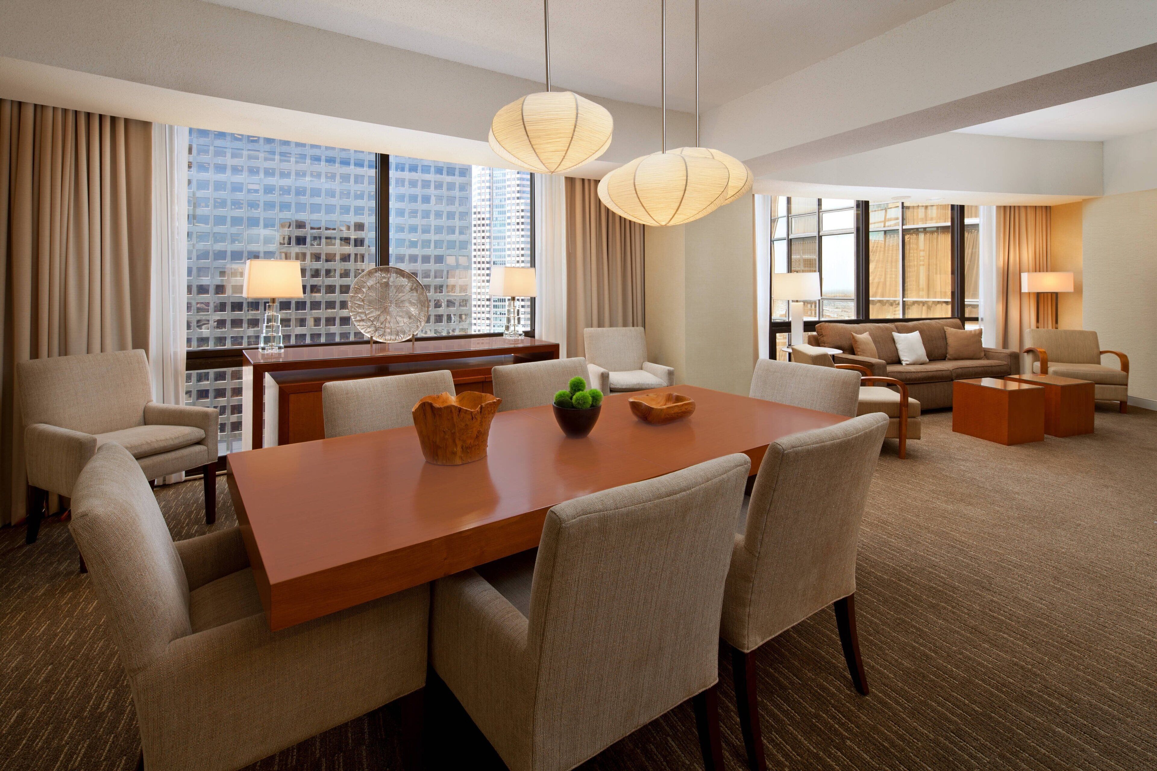 Una habitación de hotel moderna con una mesa de comedor grande, asientos cómodos y ventanas amplias que ofrecen vistas a la ciudad.