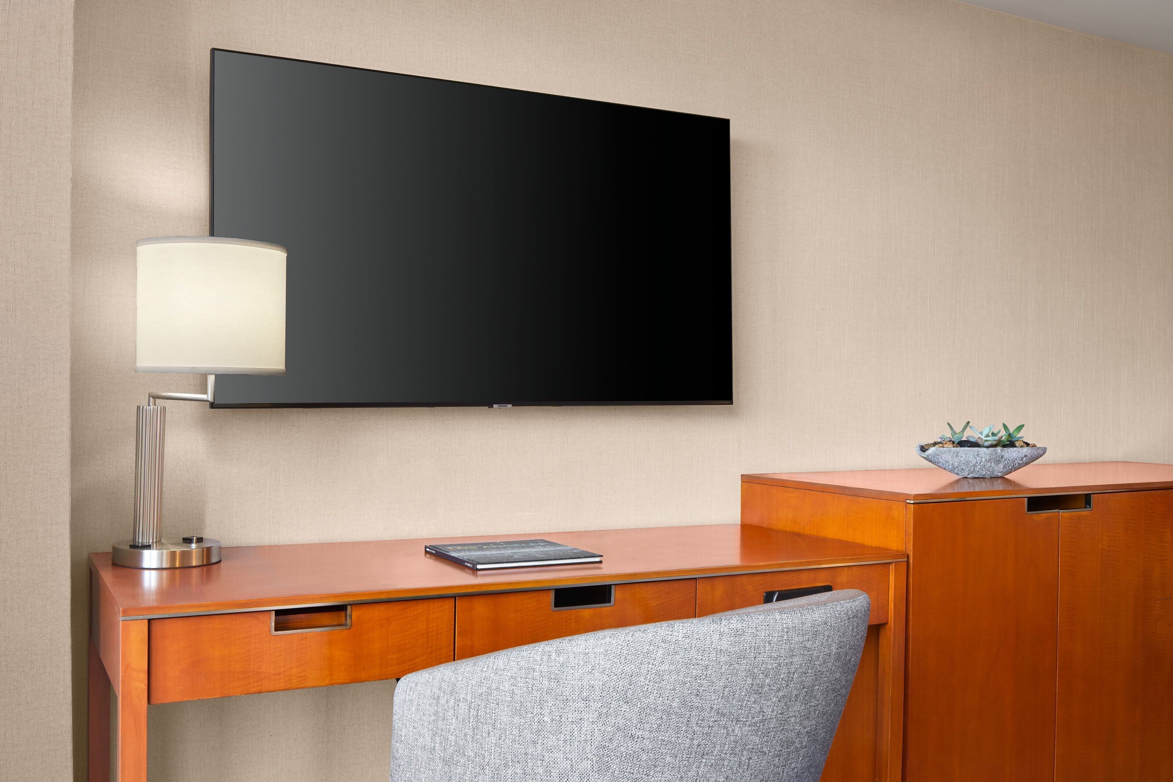 A hotel room with a wooden desk, a flat-screen TV mounted on the wall, a lamp, and a small plant on the desk.
