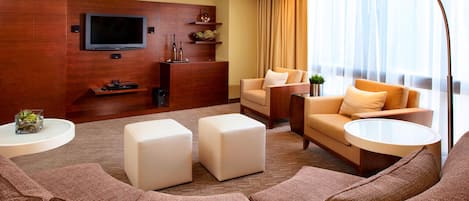 Una habitación de hotel moderna con un amplio sofá modular, un televisor de pantalla plana montado en una pared de madera, una mesita de centro y una lámpara de pie.