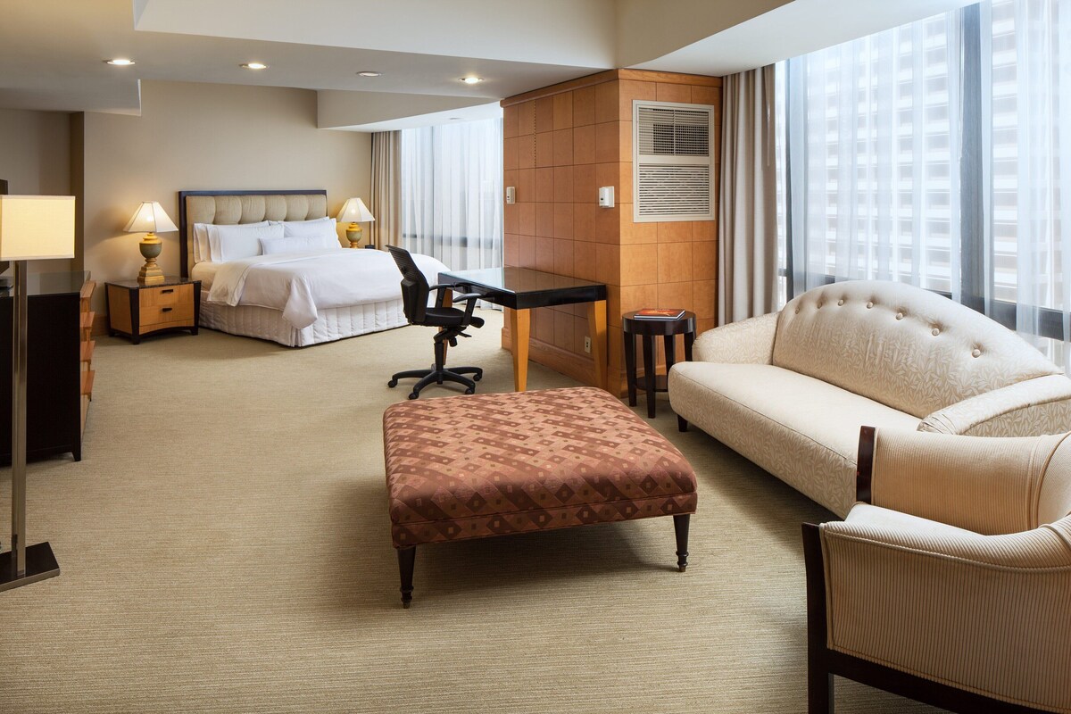 A hotel room with a bed, a desk, a chair, an ottoman, and a sofa.