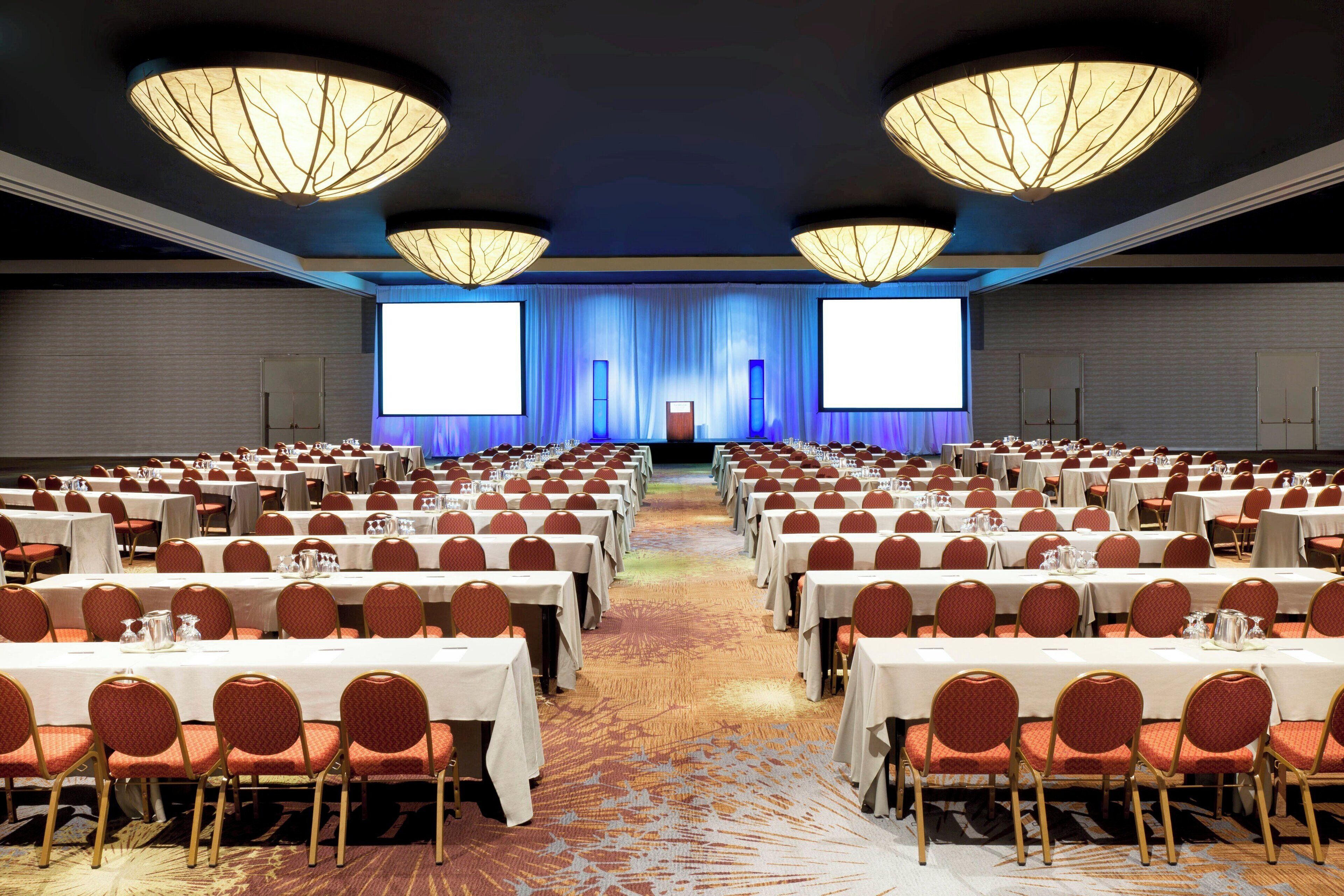 A conference room with rows of tables and chairs, a stage with a blue curtain backdrop, and large screens on either side.