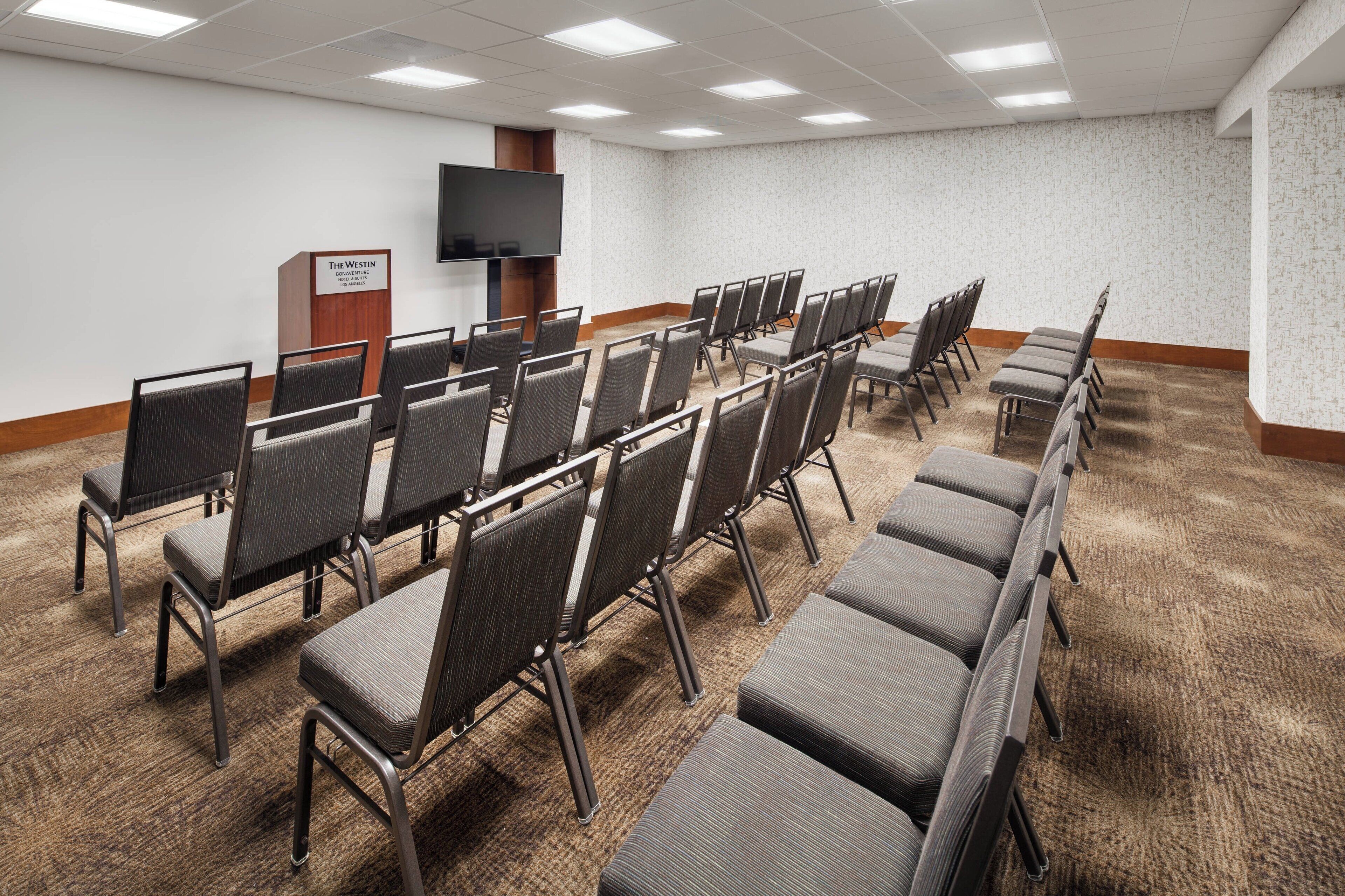 Uma sala de conferências com fileiras de cadeiras, uma TV de tela plana e um pódio de madeira.
