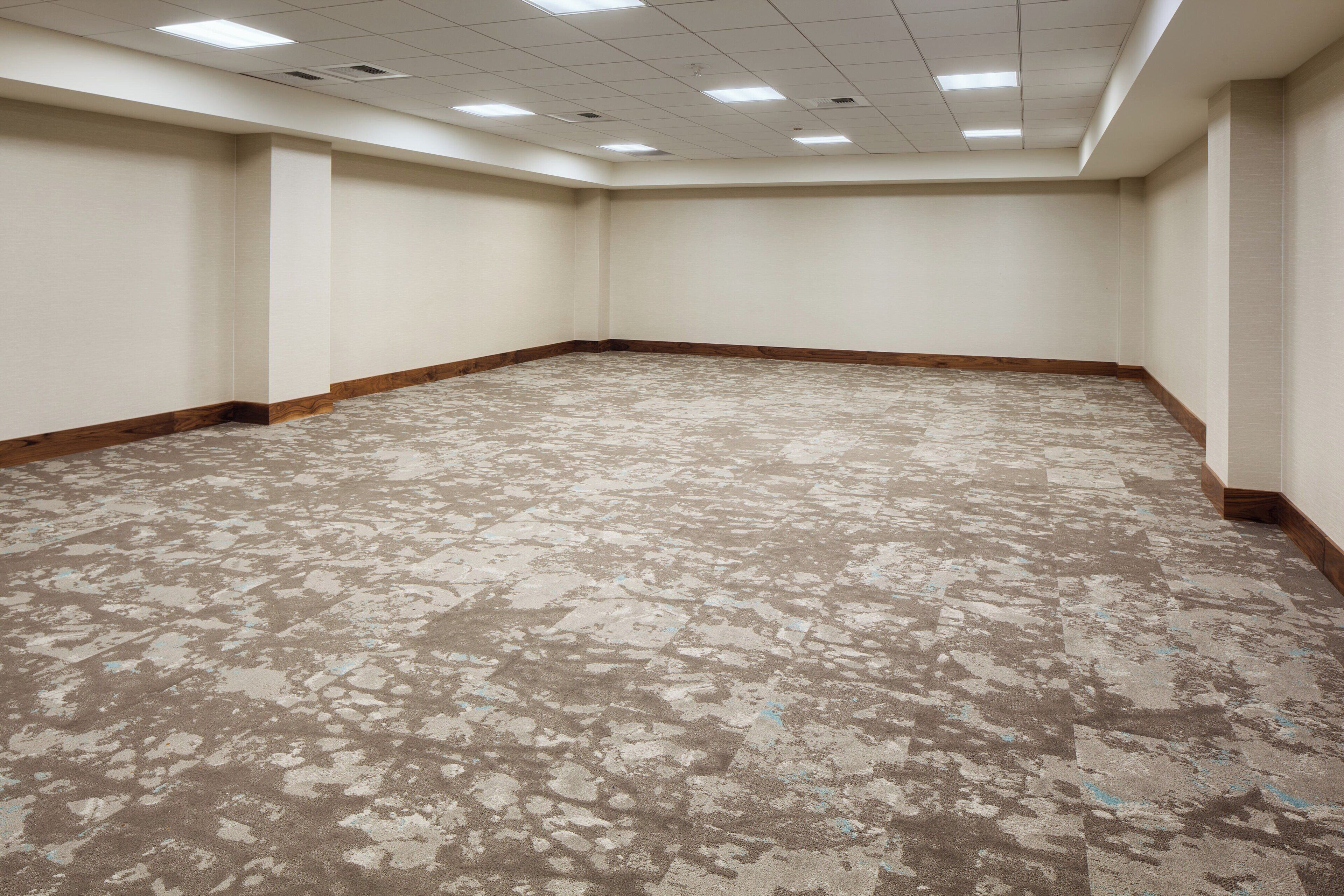 Una habitación amplia y vacía con alfombra estampada, paredes blancas y zócalo de madera.