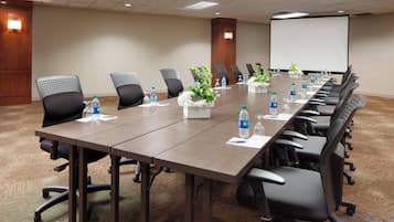 Un salón de conferencias con una larga mesa, sillas negras, botellas de agua y plantas en macetas.