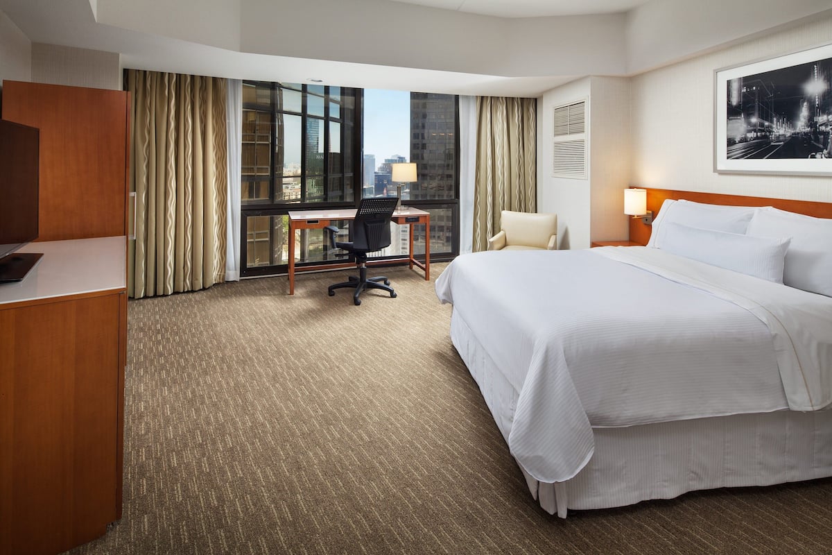 Habitación de hotel con una cama grande, un escritorio, una silla y vistas a la ciudad desde la ventana.