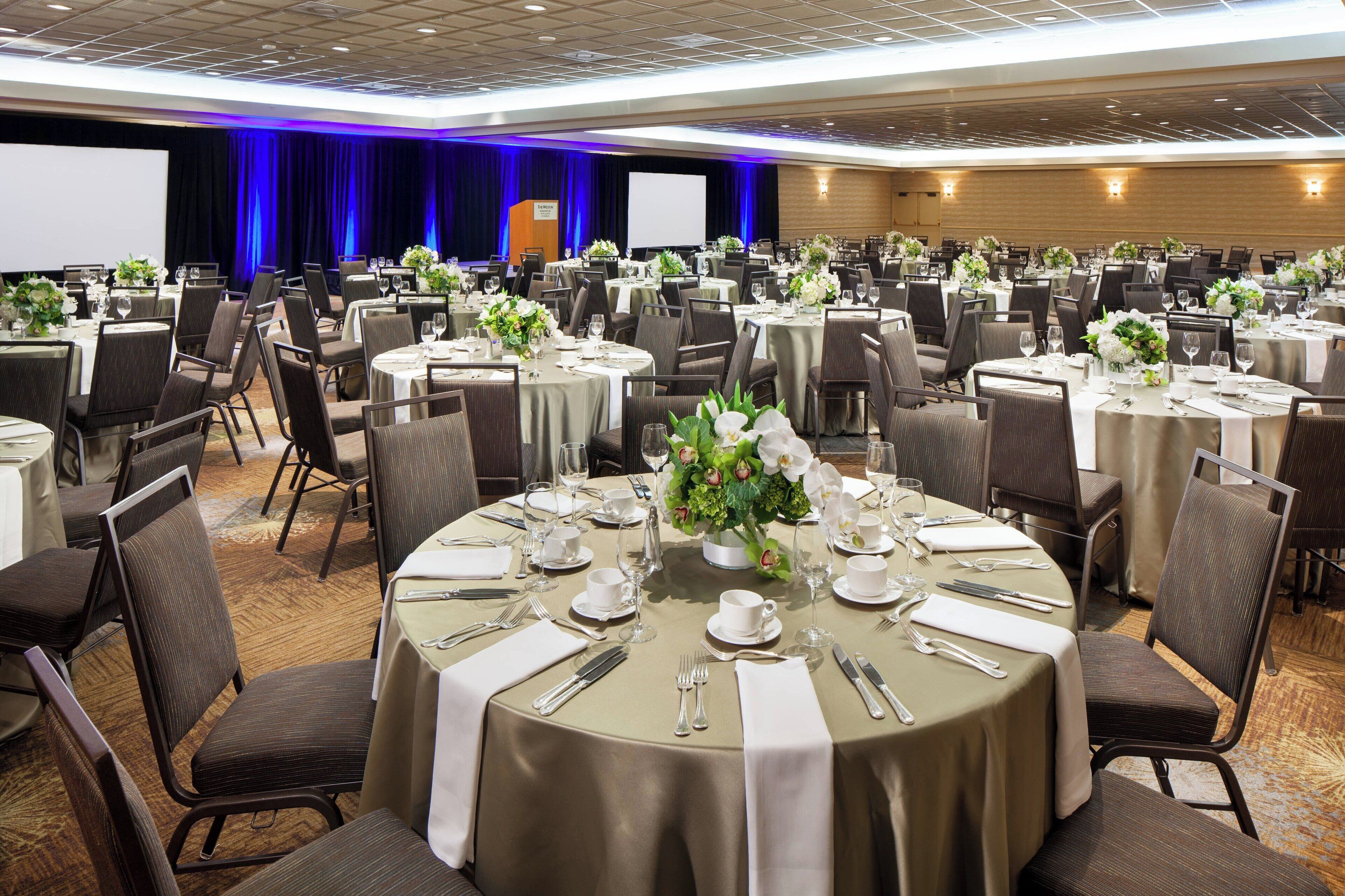 A banquet hall with round tables set for dining, chairs, and a stage with a screen.