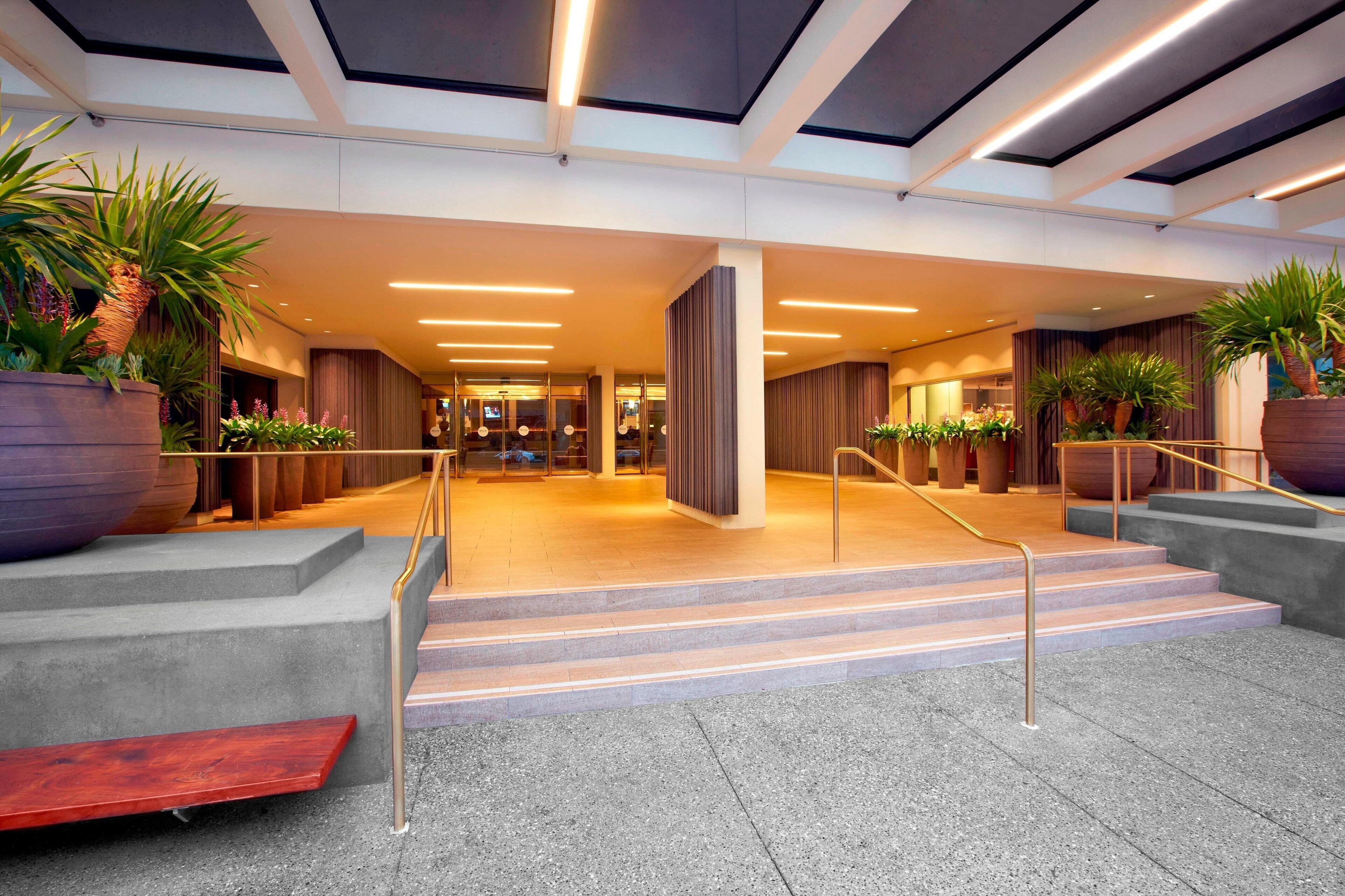 A modern hotel lobby with a spacious area, a staircase leading to the upper floor, and large potted plants.