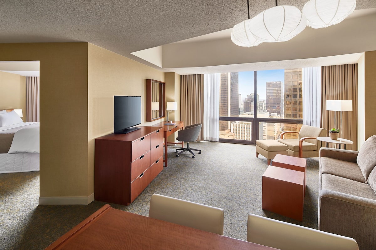 Habitación de hotel con una cama grande, televisión, escritorio, silla, sofá y vistas a la ciudad a través de amplios ventanales.