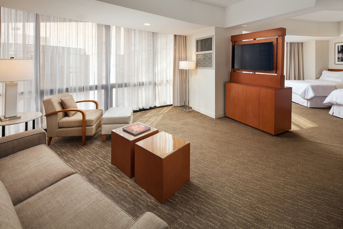 A hotel room with a sofa, armchair, coffee table, TV, and a bed.