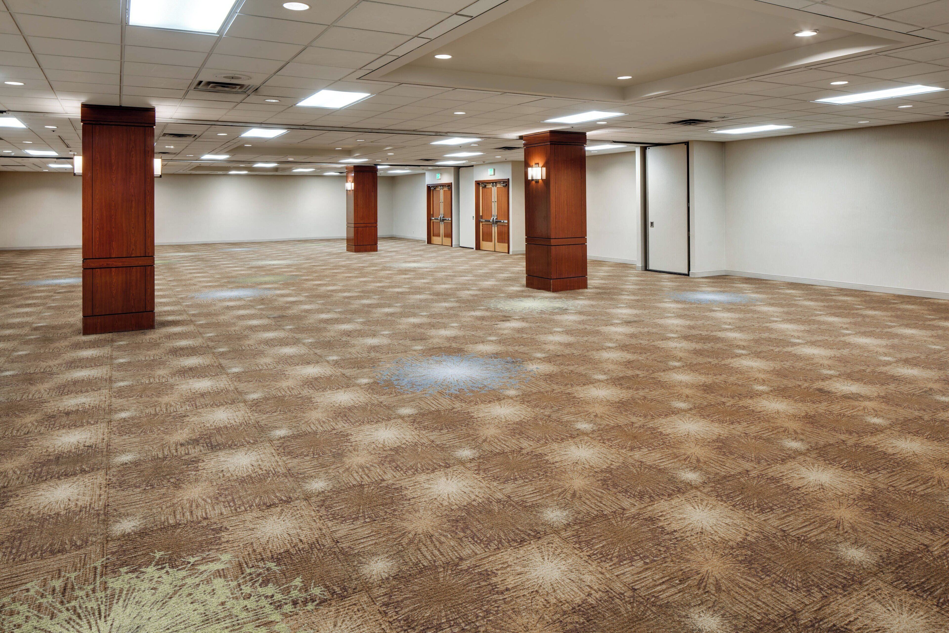 Un amplio salón de conferencias vacío con piso alfombrado, pilares de madera e iluminación empotrada.