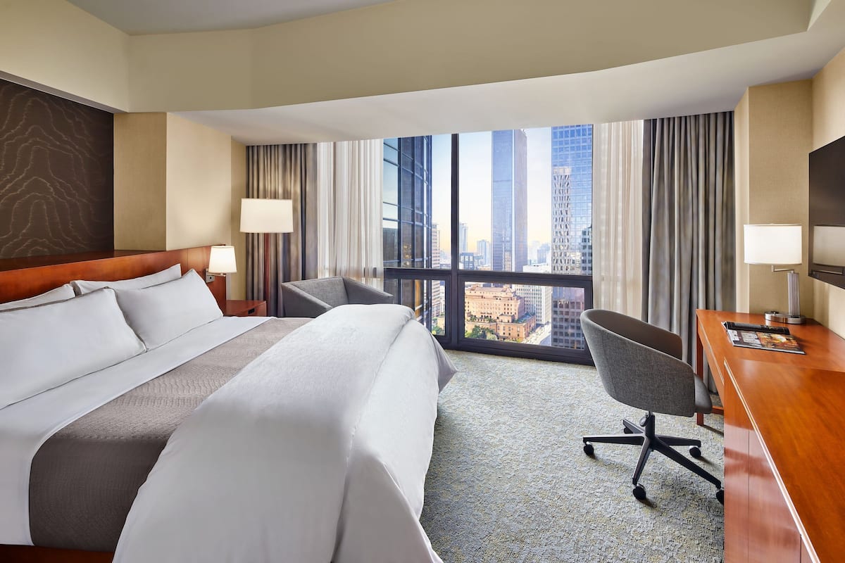 Habitación de hotel con una cama grande, un escritorio, una silla y vistas a la ciudad a través de amplios ventanales.
