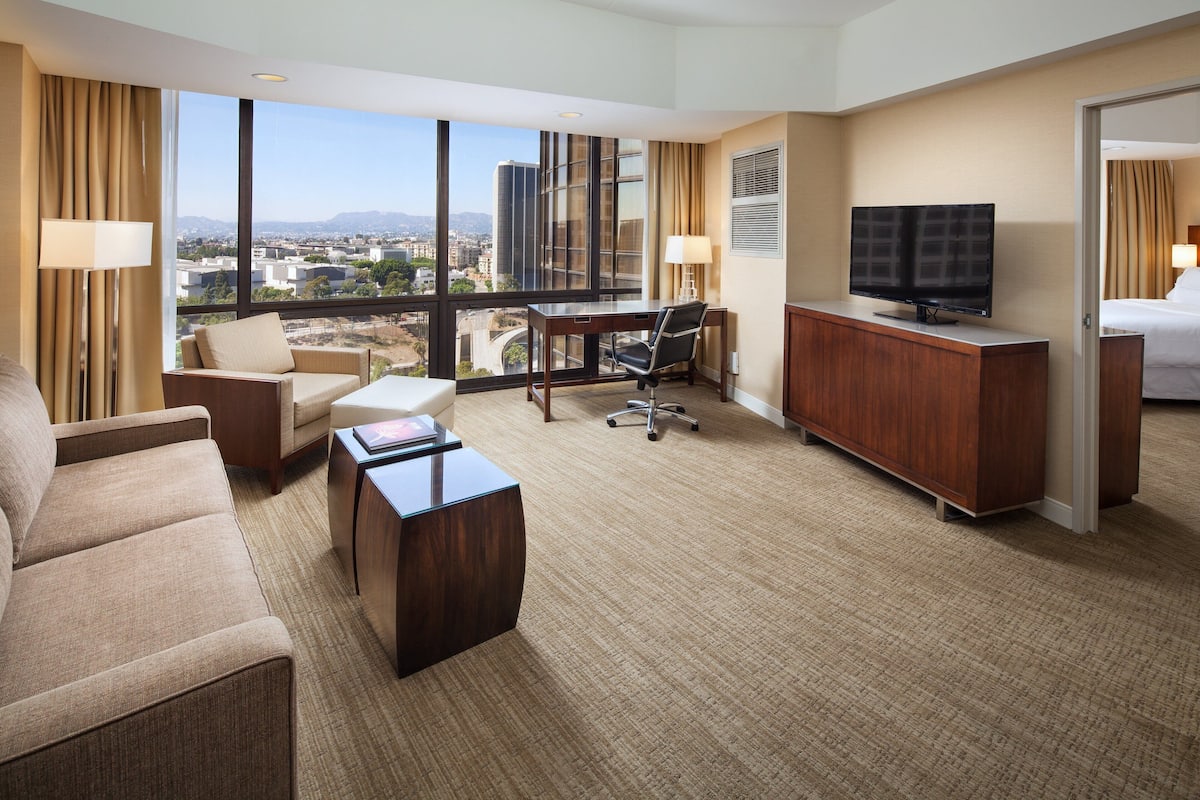 Habitación de hotel con un ventanal grande, un escritorio, televisor, cama y sofá.