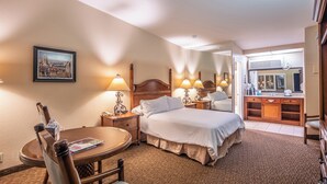A hotel room with a bed, two bedside tables with lamps, a chair, a round table, a painting on the wall, and a bathroom visible through an open door. - Casa Ojai Inn (Ojai)