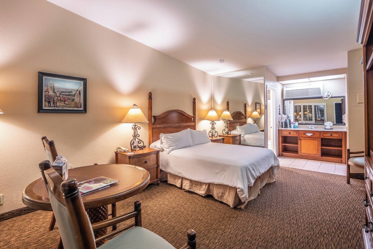Quarto de hotel com cama, duas mesas de cabeceira com luminárias, uma cadeira, uma mesa redonda, um quadro na parede e um banheiro visível através de uma porta aberta.