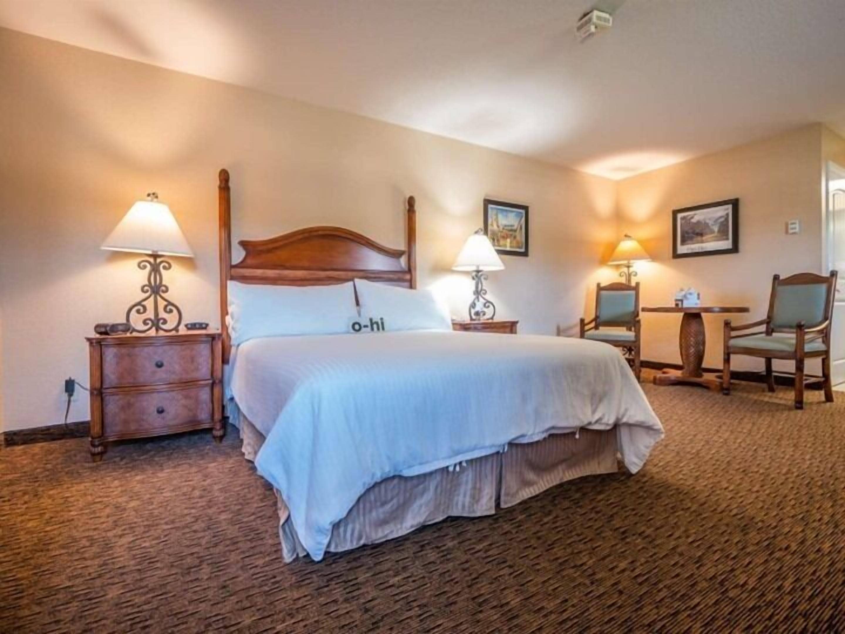 A hotel room with a large bed, two bedside tables with lamps, a wooden chair, a small table, and a framed picture on the wall.