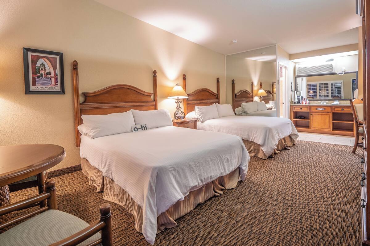 Quarto de hotel com duas camas, cabeceira de madeira, uma mesinha, uma cadeira, um espelho e um quadro na parede.