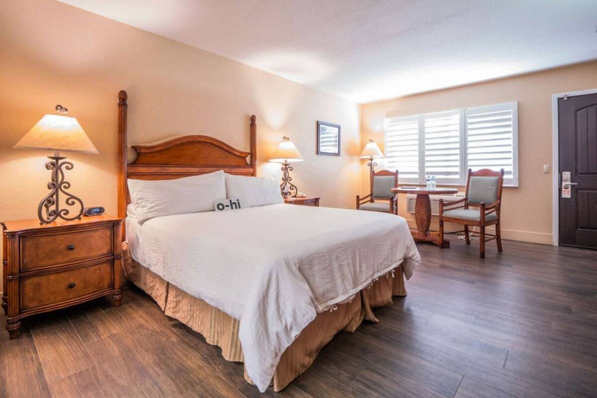 Quarto de hotel com uma cama grande, duas mesas de cabeceira com luminárias, uma cadeira de madeira, uma mesa pequena e uma janela com persianas.