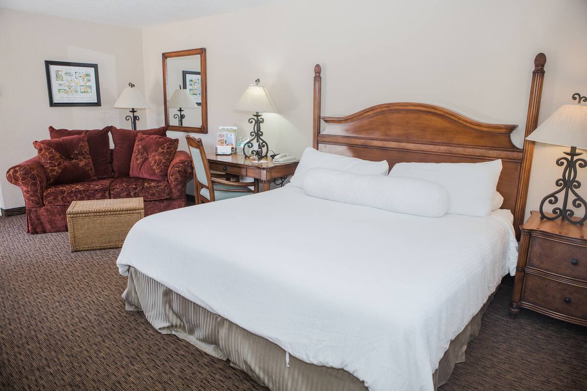 Quarto de hotel com uma cama grande, cabeceira de madeira, sofá vermelho, duas mesas de cabeceira com luminárias e um quadro pendurado na parede.