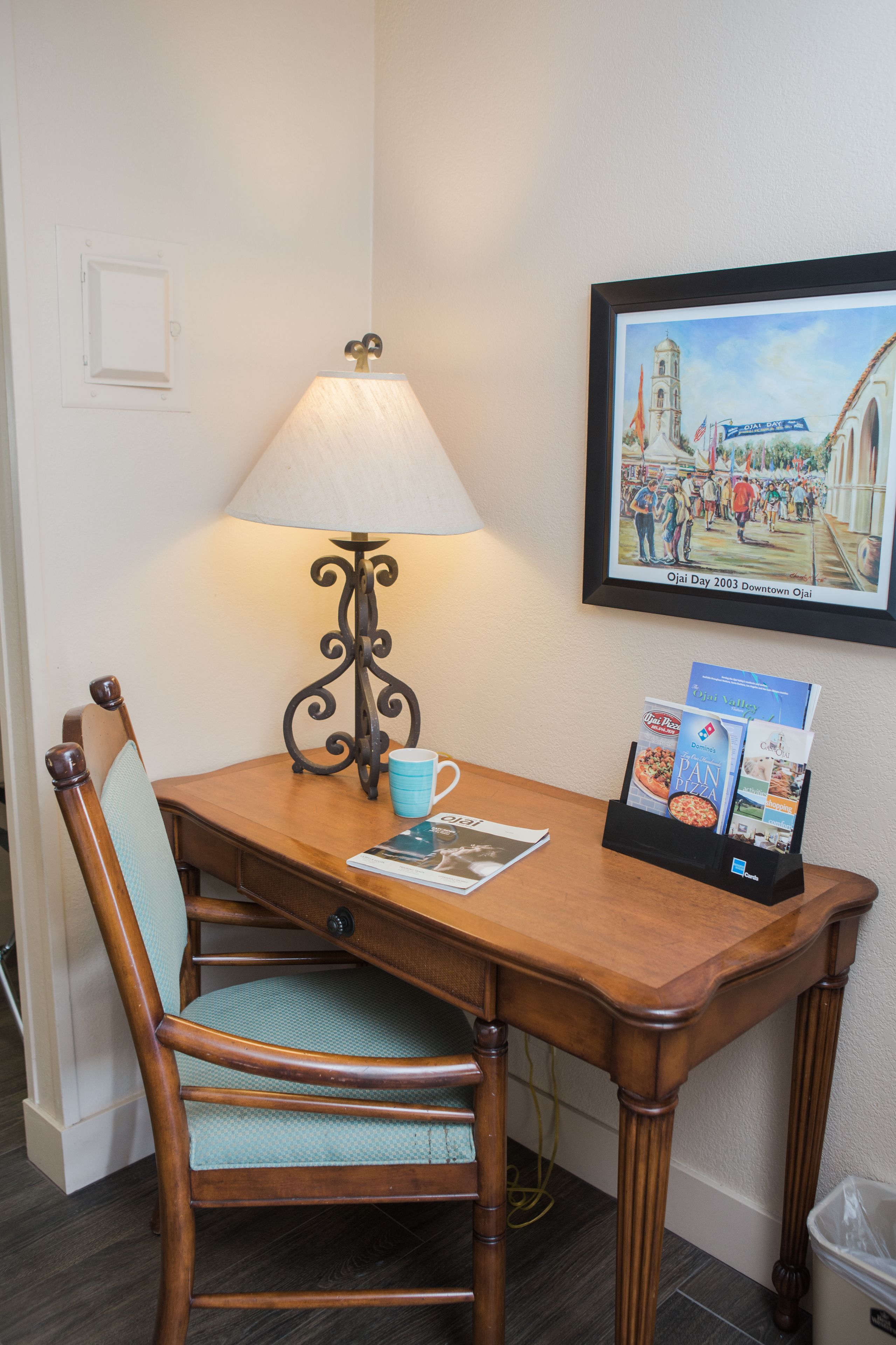 A wooden table with a lamp, a chair, a framed picture, and a magazine rack.