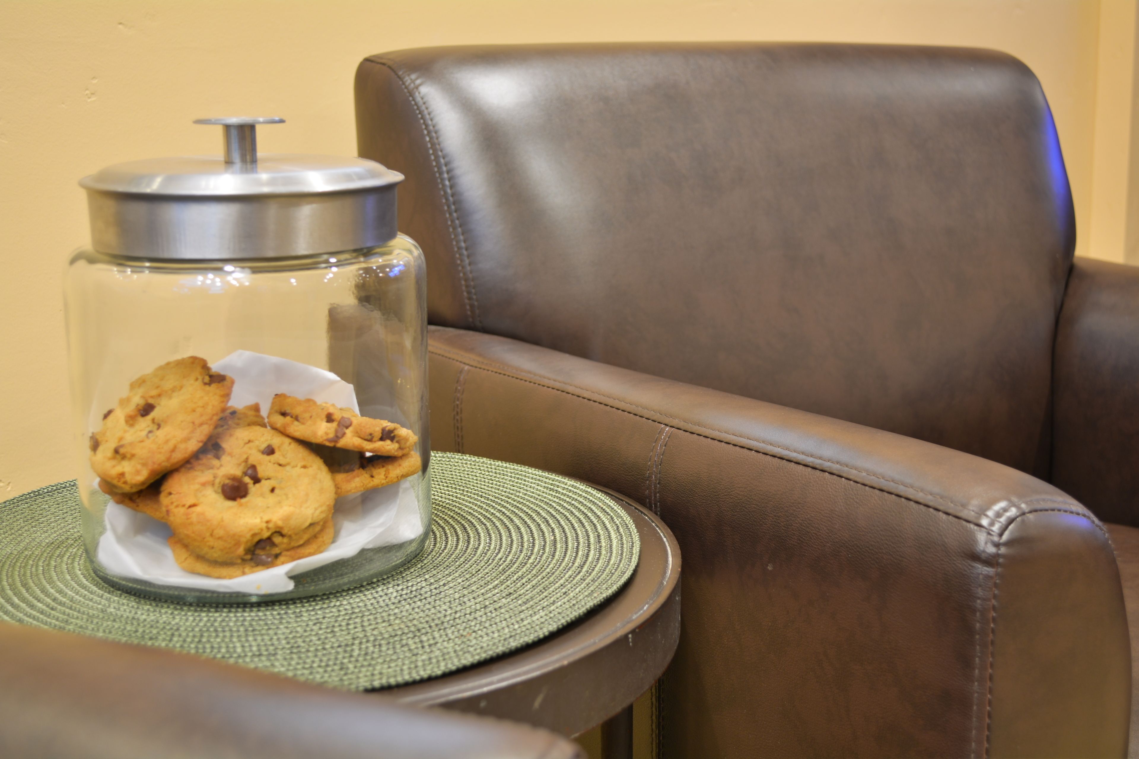 Een glazen pot met koekjes op een geweven onderzetter op een tafel naast een leren stoel.