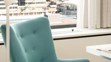 Ein saphirfarbener Sessel an einem Fenster mit Blick auf eine Stadtlandschaft, eine weiße Stehlampe und ein kleiner Tisch mit einer Zeitschrift.